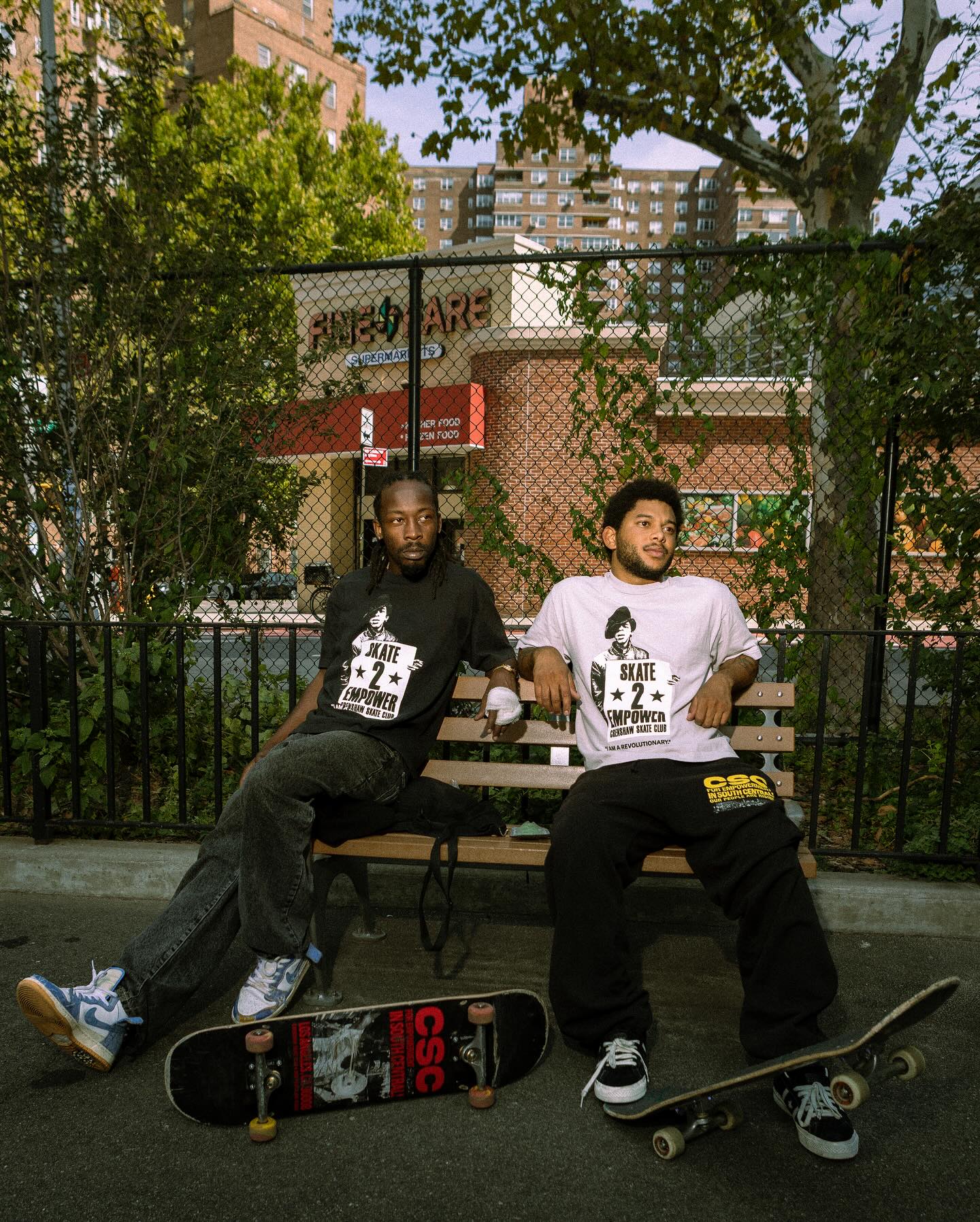 Two young men on a bench in the park with their skateboards rocking crenshaw skate club gear